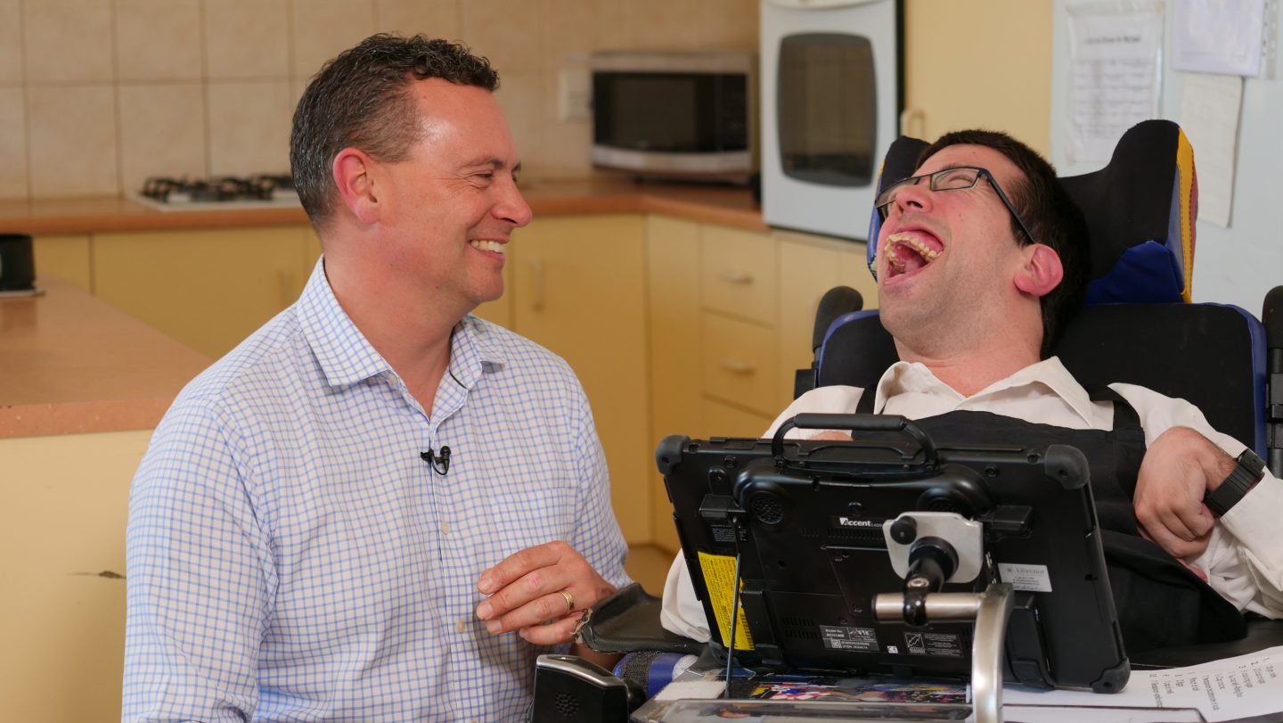 Todd is looking at Anton and smiling, and Anton is laughing. Anton uses a wheelchair.